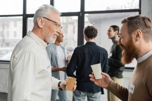 Усміхнені чоловіки з паперовими чашками розмовляють під час зустрічі алкоголіків у центрі відновлення — стокове фото