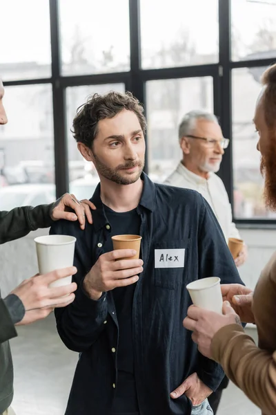 People with paper cups talking to man during alcoholics meeting in rehab center — Stock Photo