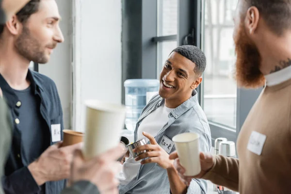 Cheerful african american man holding thermos and talking to people during alcoholics meeting in rehab center — Stock Photo
