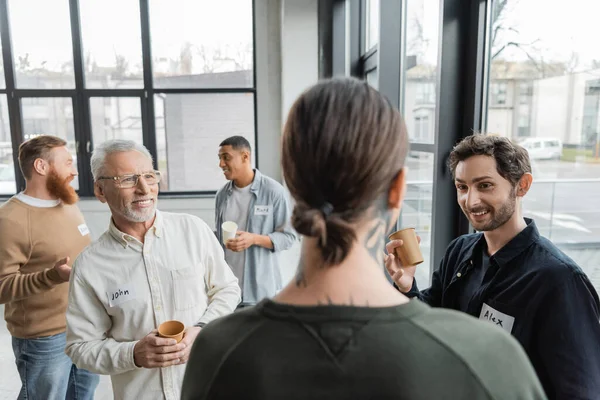 Positive people with paper cups looking at blurred tattooed man during alcoholics meeting in rehab center — Stock Photo