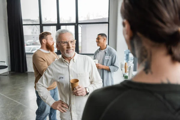 Smiling mature man with paper cup and name sticker spending time during alcoholics meeting in rehab center — Stock Photo