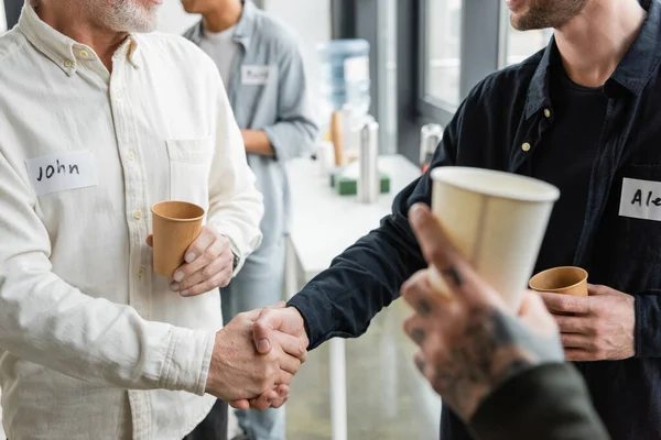 Cropped view of people holding paper cups and shaking hands during alcoholics meeting in rehab center — Stock Photo