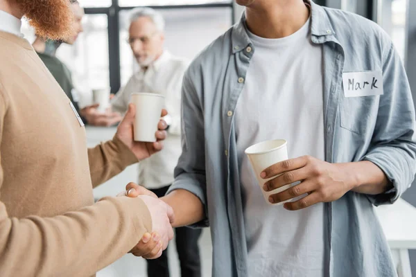 Interracial men with paper cups shaking hands during alcoholics meeting in rehab center — Stock Photo