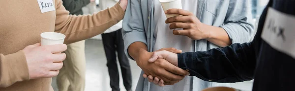Vista recortada de personas multiétnicas con adicción al alcohol sosteniendo vasos de papel y estrechando las manos en el centro de rehabilitación, pancarta - foto de stock