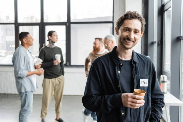 Smiling man holding paper cup and looking at camera during alcoholics meeting in rehab center — Stock Photo