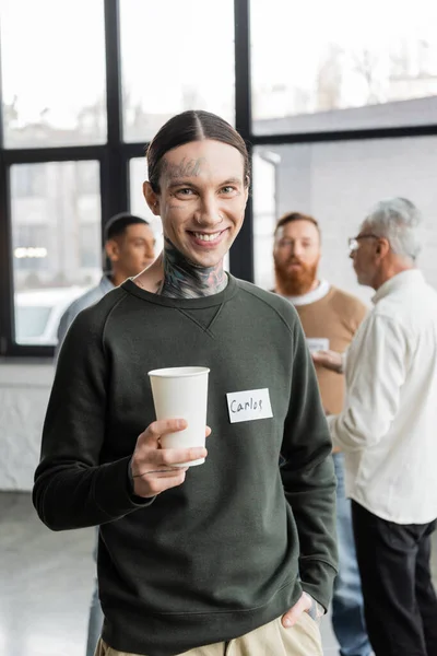 Улыбающийся татуированный мужчина с бумажной чашкой и смотрящий в камеру во время встречи алкоголиков в реабилитационном центре — стоковое фото
