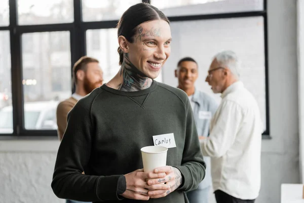 Positive tattooed man holding paper cup during meeting of alcoholics in recovery center — Stock Photo