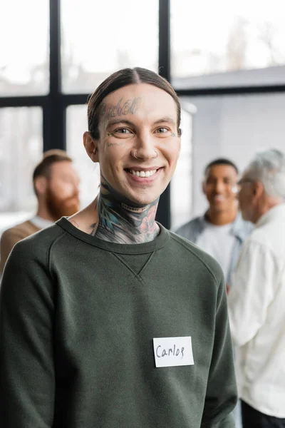 Retrato del hombre tatuado sonriente con el nombre pegatina mirando a la cámara durante una reunión aa en el centro de rehabilitación - foto de stock