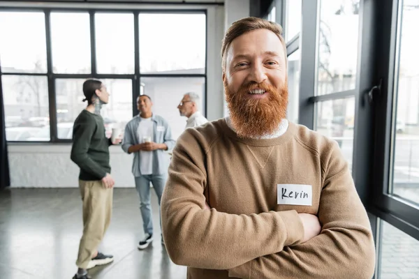 Cheerful bearded man crossing arms and looking at camera during alcoholics meeting in rehab center — Stock Photo