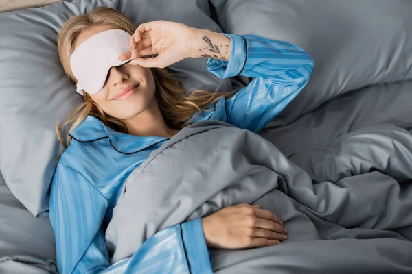 Вид зверху щаслива блондинка у сплячій масці та шовковій піжамі, що відпочиває у ліжку — стокове фото