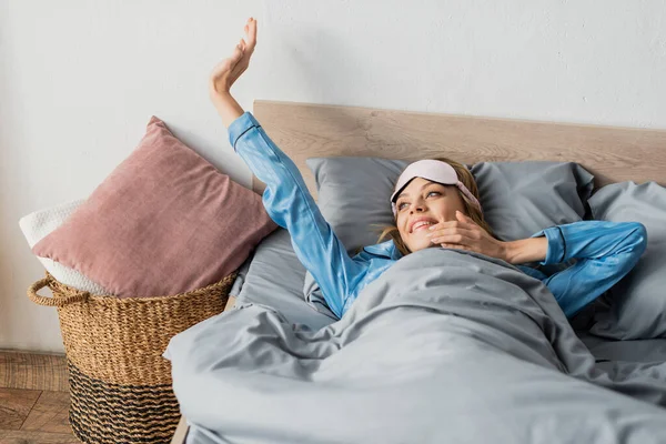 Весёлая женщина в маске для сна и ночной одежде, растягивающаяся в постели — стоковое фото