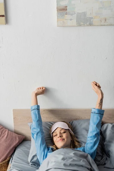 Mujer tatuada con máscara para dormir y ropa de dormir estirándose mientras está acostada en la cama - foto de stock
