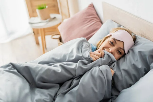 Glückliche junge Frau in Schlafmaske liegt unter Decke in bequemem Bett — Stockfoto