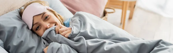 Счастливая молодая женщина в маске для сна лежит под одеялом в удобной постели, баннер — стоковое фото