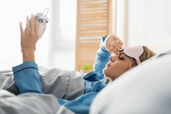 Side view of young woman in sleeping mask looking at alarm clock while lying on bed — Stock Photo