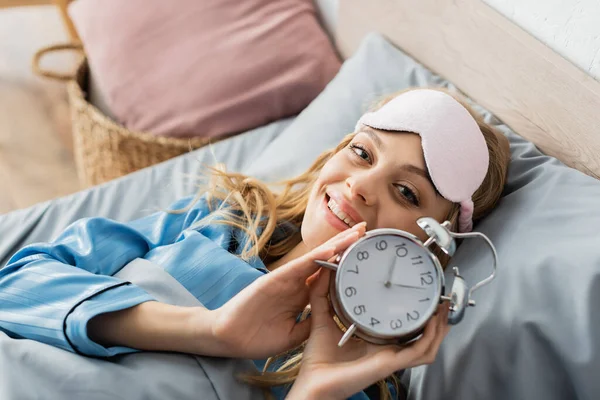 Vista superior de la mujer feliz en la máscara de dormir y pijama azul sosteniendo el reloj despertador mientras está acostado en la cama - foto de stock