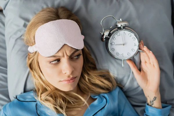 Vista superior da mulher tatuada em máscara de dormir e pijama azul olhando para o despertador enquanto deitado na cama — Fotografia de Stock