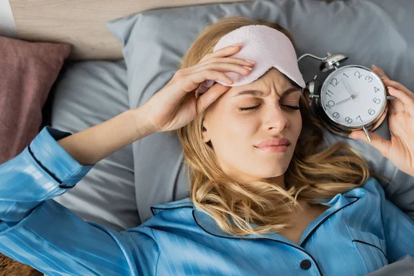Top view of displeased woman in sleeping mask and blue pajama holding alarm clock while lying in bed — Stock Photo