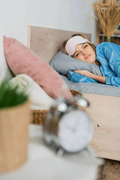 Mulher descontente em máscara de dormir e pijama deitado na cama perto de despertador desfocado — Fotografia de Stock