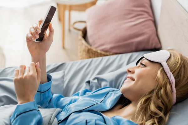 Fröhliche Frau in Schlafmaske und blauem Pyjama mit Smartphone im Bett — Stockfoto