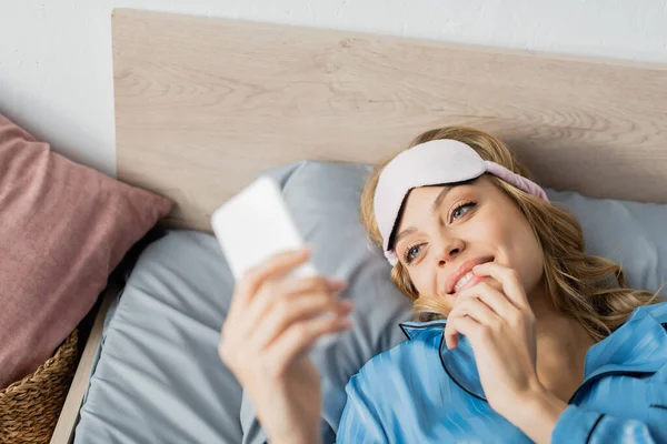Vista de ángulo alto de la mujer alegre en la máscara de dormir y el pijama azul con el teléfono inteligente en la cama - foto de stock