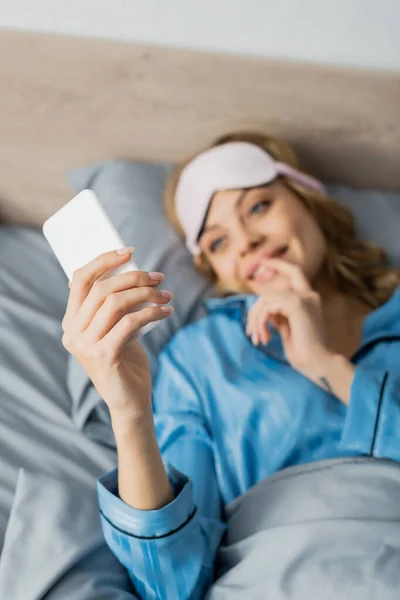 Mujer borrosa y alegre con máscara para dormir y pijama azul usando smartphone en la cama - foto de stock