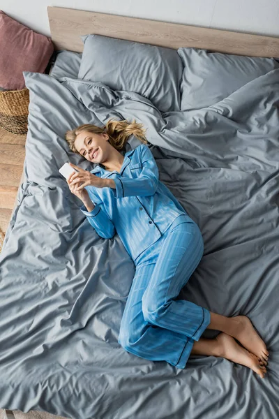 Top view of cheerful woman in blue pajama using smartphone in bed — Stock Photo