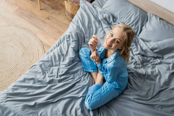 Вид сверху положительной женщины в синей пижаме, смотрящей в камеру сидя на кровати — стоковое фото