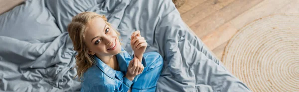 Positive Frau im blauen Pyjama, die im Sitzen auf dem Bett in die Kamera blickt, Banner — Stockfoto