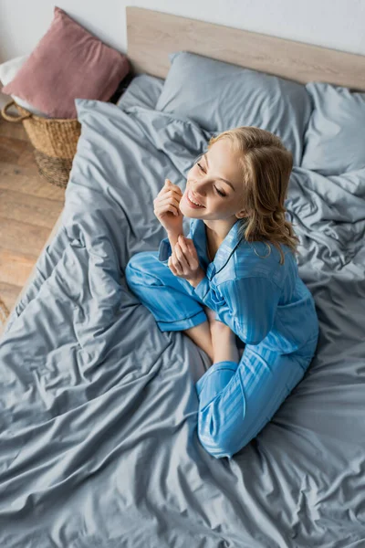 Draufsicht auf eine fröhliche junge Frau im blauen Pyjama, die zu Hause auf dem Bett sitzt — Stockfoto