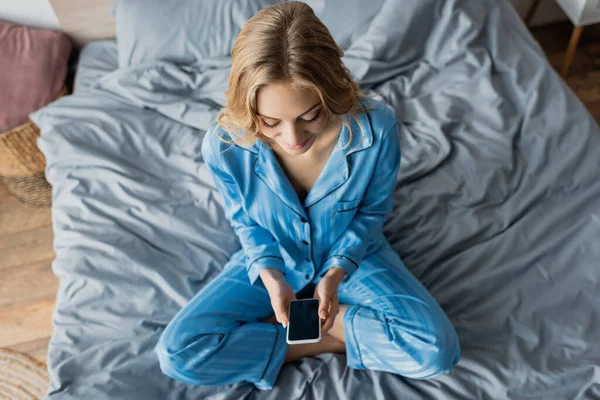 Vista dall'alto di donna allegra in pigiama blu utilizzando smartphone con schermo bianco mentre si siede sul letto — Foto stock
