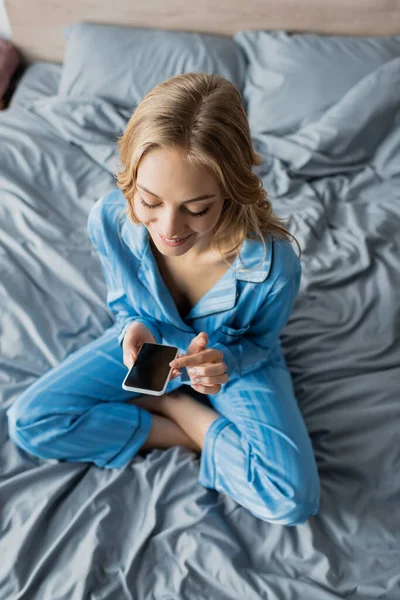 Vue du haut de la femme souriante en pyjama bleu à l'aide d'un smartphone avec écran blanc assis sur le lit — Photo de stock