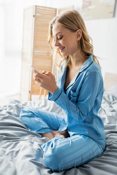 Веселая женщина в синей пижаме с помощью мобильного телефона в спальне — стоковое фото