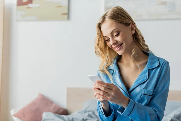 Mulher satisfeita em pijama azul usando telefone celular no quarto — Fotografia de Stock
