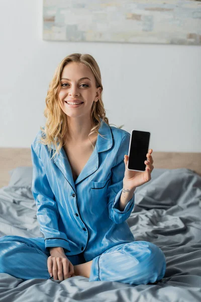 Femme satisfaite en pyjama bleu tenant smartphone avec écran blanc — Photo de stock