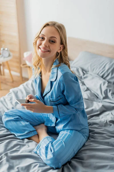 Femme positive en pyjama bleu tenant smartphone avec écran blanc — Photo de stock