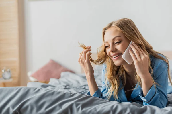 Mujer alegre y rubia hablando en el teléfono inteligente mientras está acostado en la cama en el apartamento moderno - foto de stock