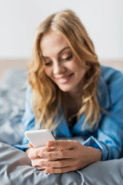 Fröhliche Frau, die im blauen Pyjama auf dem Bett liegt und Nachrichten auf dem Smartphone sendet — Stockfoto