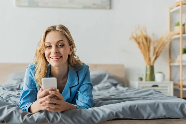 Радісна молода жінка обмінюється повідомленнями на смартфоні, лежачи в синій піжамі на ліжку — стокове фото