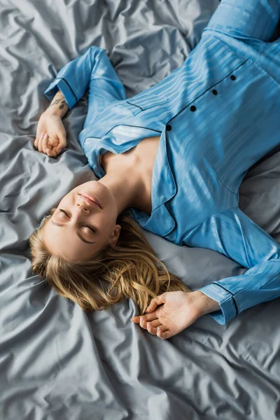 Вид сверху женщины в голубой шелковой пижаме, лежащей с закрытыми глазами на кровати в современной квартире — стоковое фото