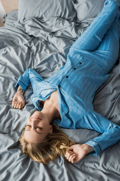 Vista superior de la mujer complacida con tatuaje acostado con los ojos cerrados en la cama en el apartamento moderno - foto de stock