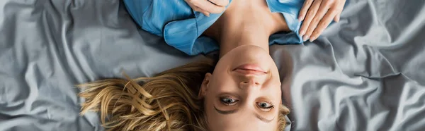 Draufsicht auf eine unbeschwerte Frau im blauen Seidenpyjama, die auf dem Bett liegt und in die Kamera blickt, Banner — Stockfoto