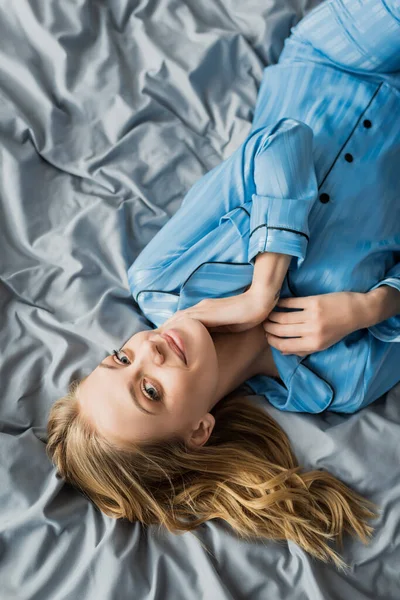 Vista superior de la mujer alegre en pijama de seda azul acostado en la cama y mirando a la cámara - foto de stock