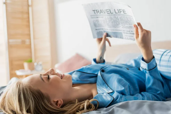 Feliz joven sonriendo mientras lee la vida de viaje periódico en la cama - foto de stock