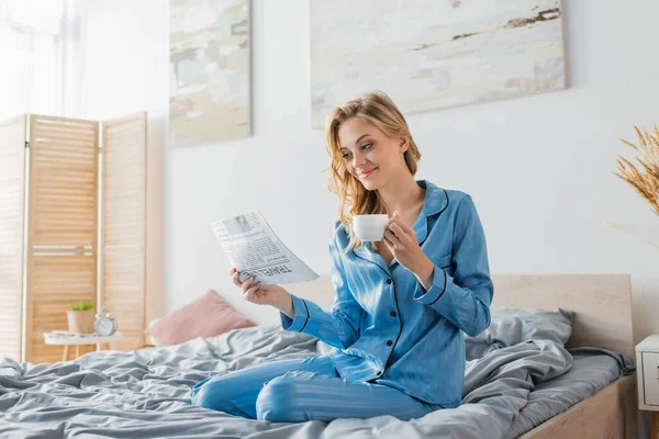 Feliz joven leyendo el periódico de viaje y sosteniendo la taza de café en la cama - foto de stock