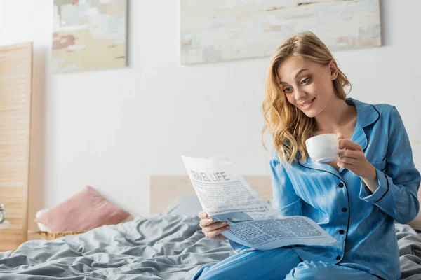 Mujer joven sorprendida leyendo el periódico de la vida de viaje y sosteniendo la taza de café en la cama - foto de stock