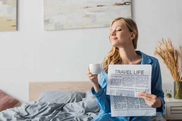 Mujer joven satisfecha sosteniendo el periódico de la vida del recorrido y la taza de café en dormitorio - foto de stock