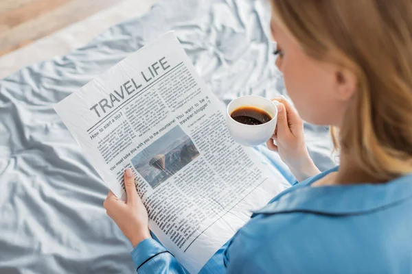 Overhead view of young woman reading travel life newspaper and holding cup of coffee in bed — Stock Photo