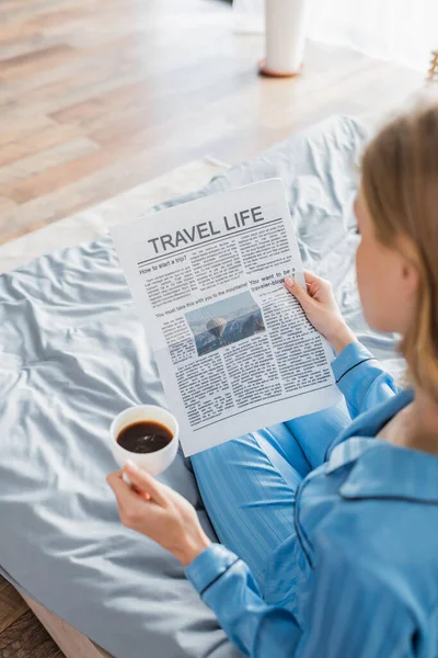 Vista aérea de la mujer rubia leyendo el periódico de la vida de viaje y sosteniendo la taza de café en el dormitorio - foto de stock