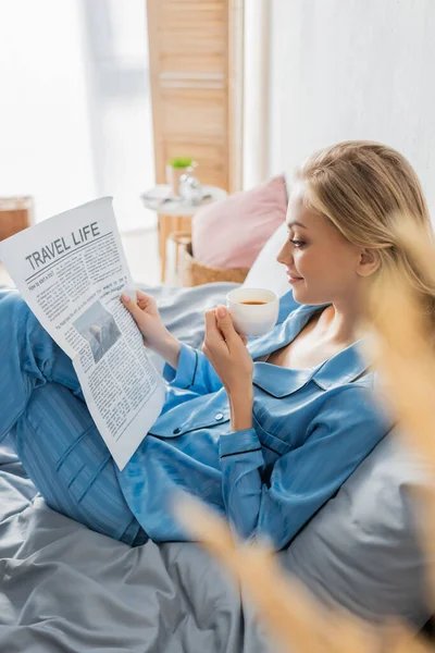 Vista lateral de la joven contenta leyendo el periódico de la vida de viaje y sosteniendo la taza de café en la cama - foto de stock